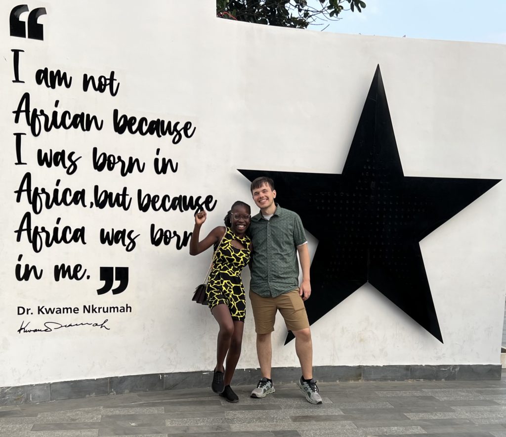 Photo of Pearl and her husband in front of a wall featuring a quote by Dr. Kwame Nkrumah, stating "I am not African because I was born in Africa, but because Africa was born in me."