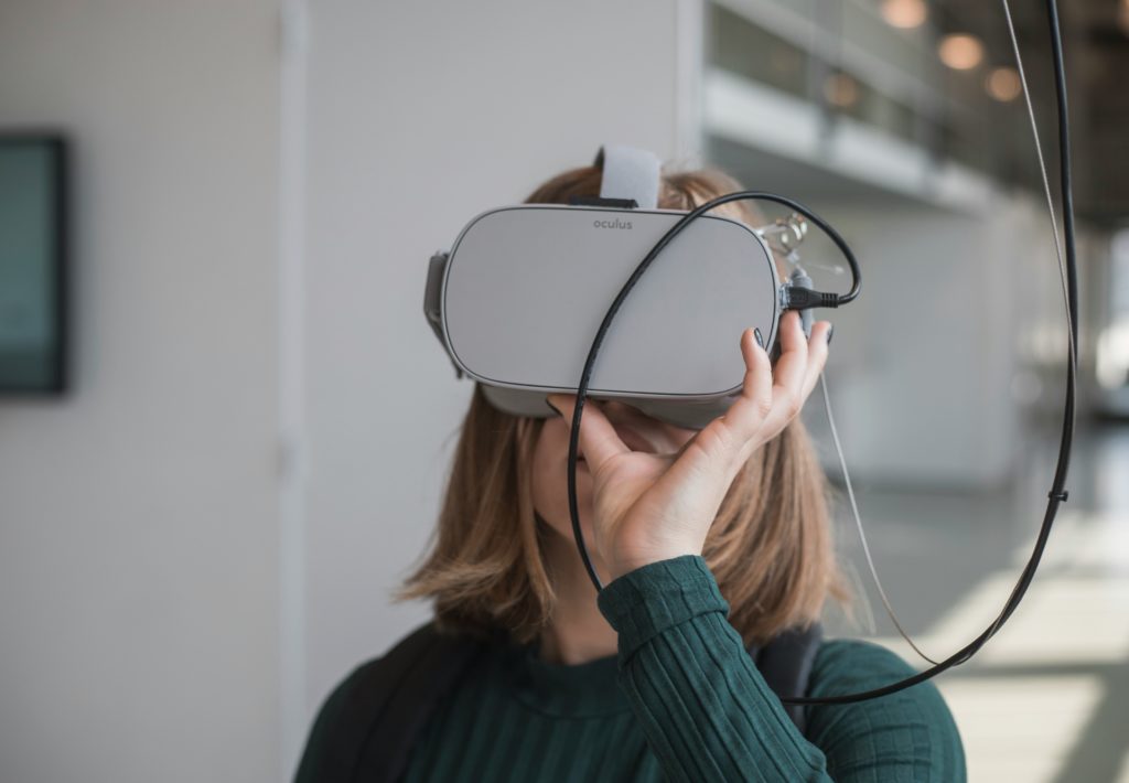 A woman holds a VR headset up to her face, while a series of cords runs down to the device from the ceiling.